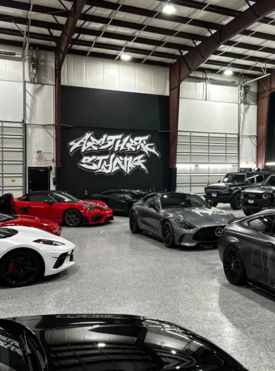 A showroom-style garage with polished flooring displays a collection of luxury sports cars, including red, white, black, and silver models. A large graffiti-style mural on the back wall reads “Aesthetic Studio.”