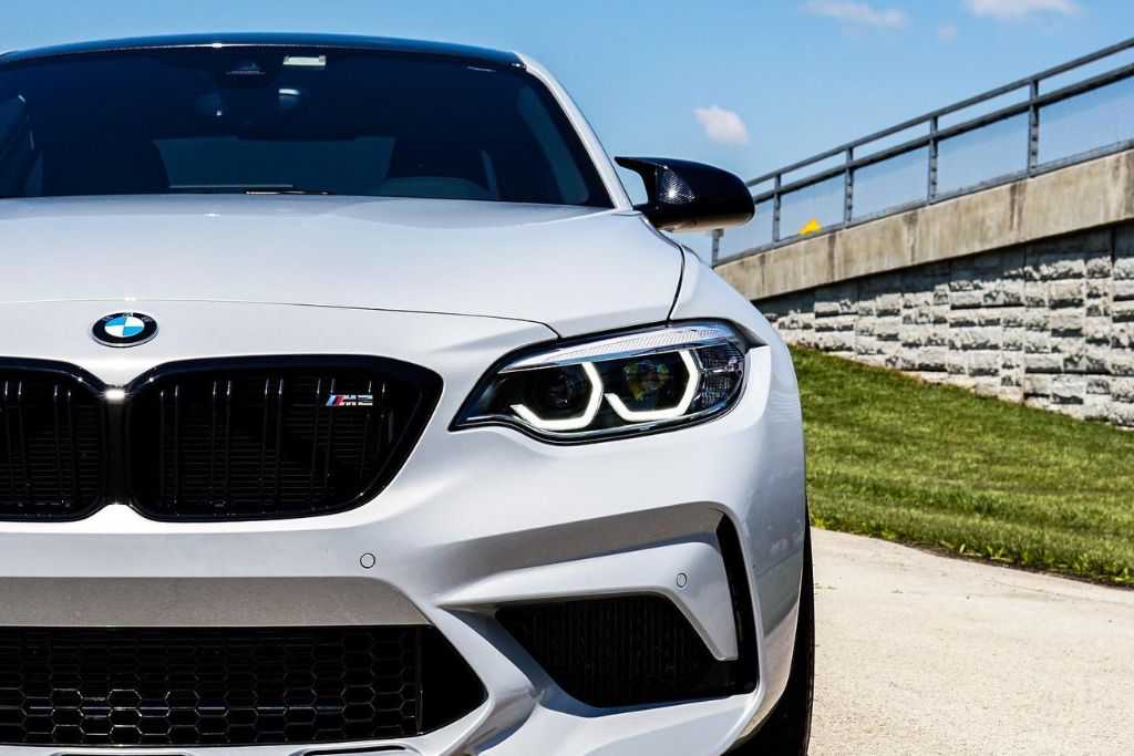 Front view of a white BMW M2 parked outdoors with sunlight highlighting its pristine paint protected by Paint Protection Film. The clear film defends against road debris, UV rays, bug splatter, and other daily driving hazards.