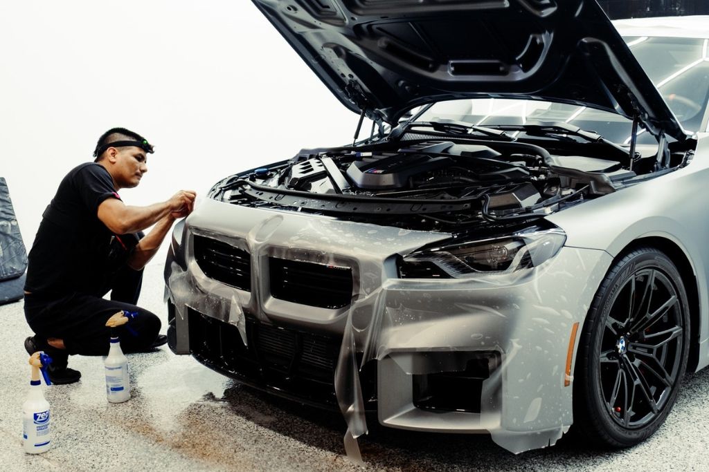 A technician carefully applies Paint Protection Film to the front of a matte silver BMW with its hood open, using tools and spray bottles for precision. The multi-layered film is visibly being smoothed onto the bumper, demonstrating how Paint Protection Film works to shield high-impact areas while preserving the car’s finish.