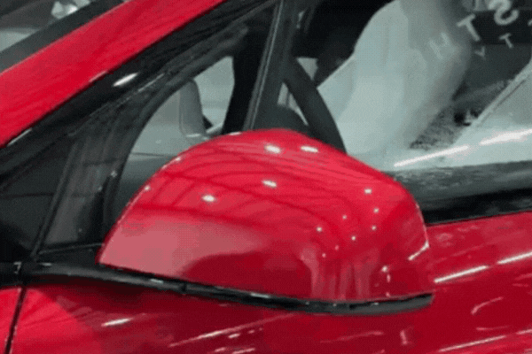 Close-up of a red car's side mirror and door area showing a smooth, glossy finish protected by Paint Protection Film. The film adds a nearly invisible layer that guards against scratches, chips, and environmental wear.