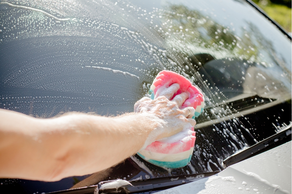 A person cleaning a car windshield with a colorful sponge and soapy water, demonstrating one of the key maintenance tips to preserve window tint longevity—using gentle, ammonia-free cleaning methods to protect the film.