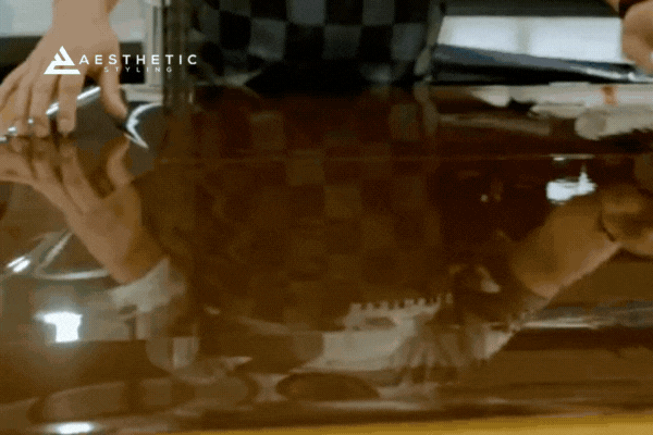 Close-up of a professional installer at Aesthetic Styling applying a dark ceramic window tint with precision, using gloved hands to smooth out the film over the car glass. The tint reflects the checkered pattern of the installer’s shirt, showing the high-gloss, bubble-free finish.