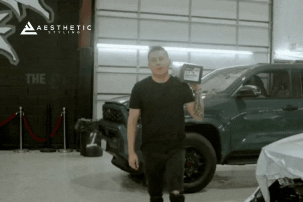 A man in a black shirt holds up a box of window tint film inside a custom auto shop, standing beside a dark green SUV with aftermarket styling. The Aesthetic Styling logo is visible in the top left corner, emphasizing the shop's branding.