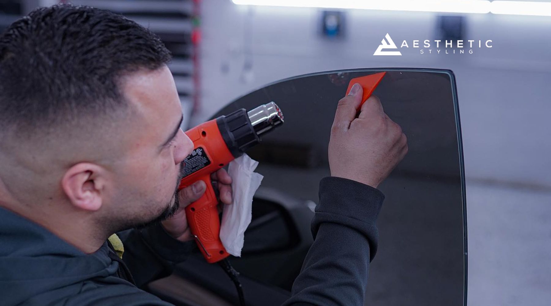 A technician in Naperville applies window tint film to a car window using a heat gun and squeegee during a professional installation. This image visually answers the question “What is window tinting” by showing the precision and tools involved in the process.