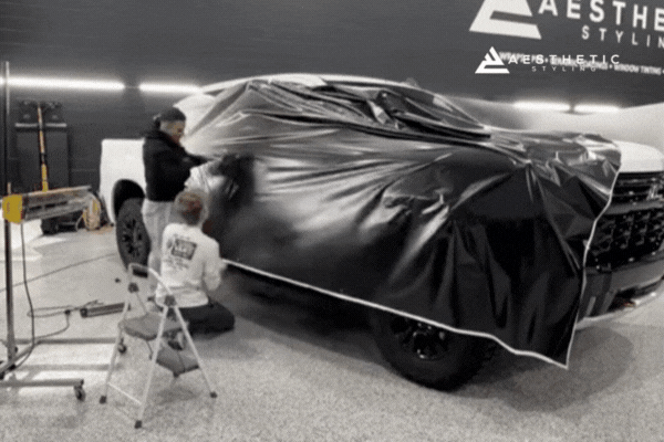 Two installers at Aesthetic Styling apply a large sheet of black vinyl wrap to the side of a white truck in a well-lit shop, carefully smoothing the material across the surface.