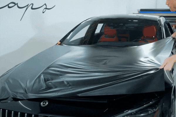 Two installers stretch a large sheet of matte black vinyl wrap across the hood of a black luxury car with red interior, preparing to smooth it over the surface.