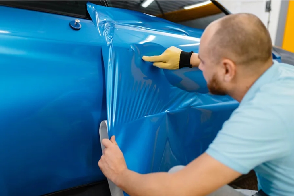 A man wearing a glove applies a vibrant blue vinyl wrap to the side panel of a car, carefully smoothing the material over the surface with his hand.
