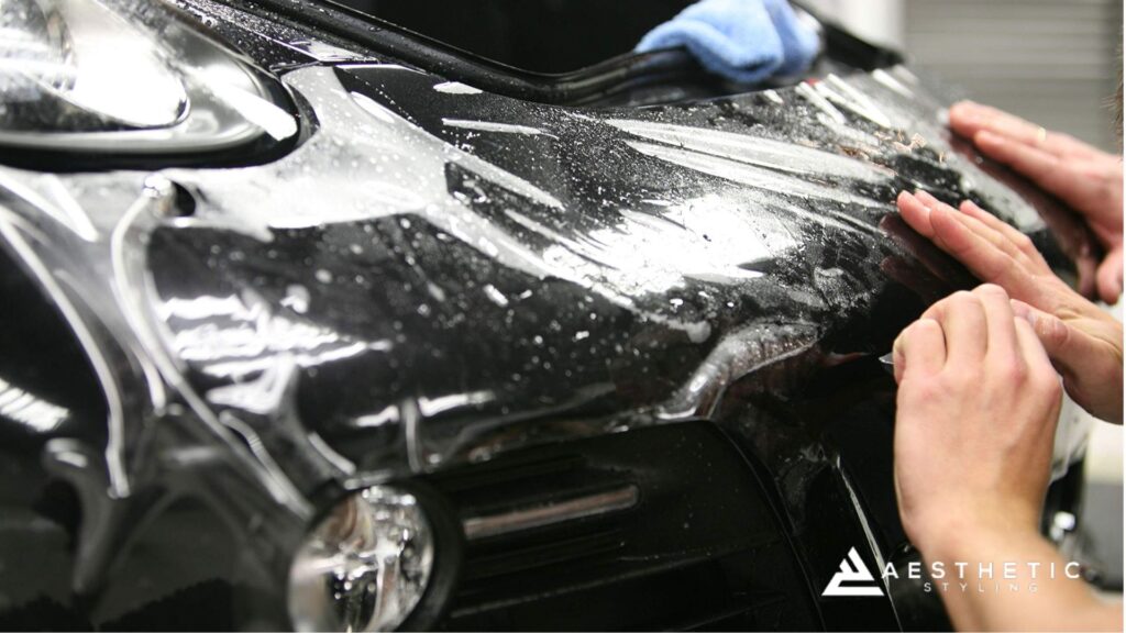 Two hands pressing and smoothing paint protection film onto the front bumper of a black car, with water droplets visible on the surface.