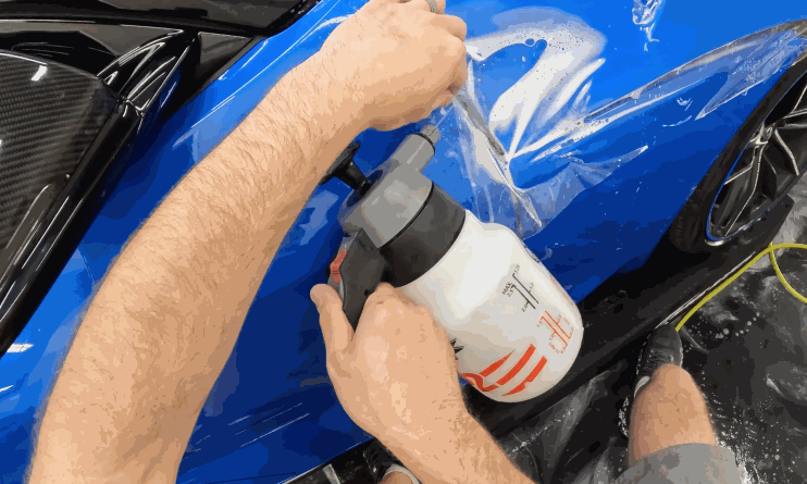 Close-up of a person applying paint protection film (PPF) to a blue car fender using a pressure sprayer and smoothing the film with their hands.