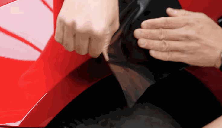 Hands stretch and press a black vinyl wrap over the curved edge of a red car, ensuring it adheres tightly to the surface.