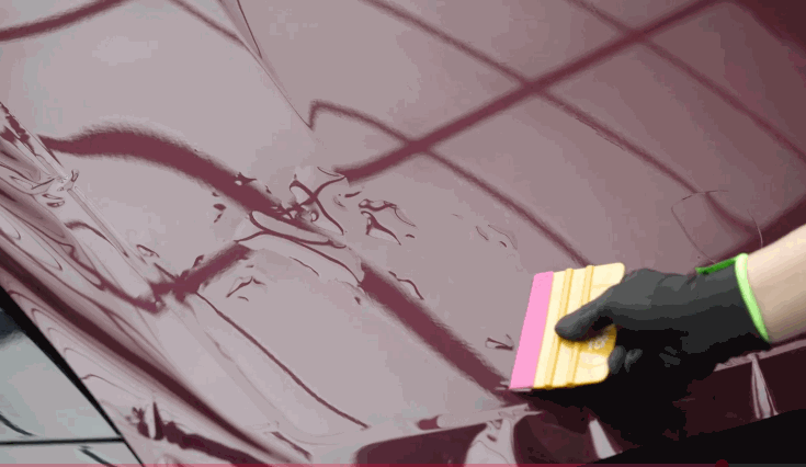 A gloved hand uses a squeegee to smooth a deep burgundy vinyl wrap onto a car's hood, eliminating air bubbles for a clean finish.