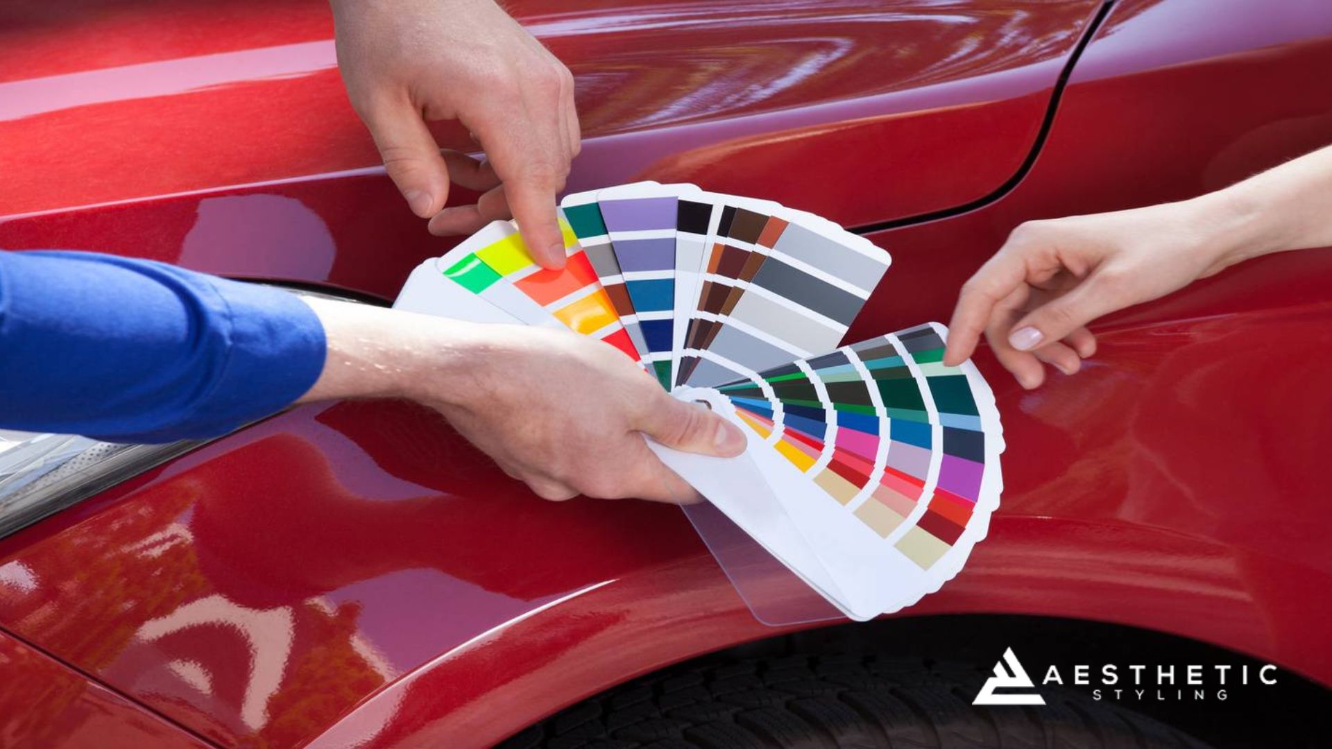 Two people hold a fan of color swatches next to a red car to compare and select from a wide range of vinyl or paint finish options.