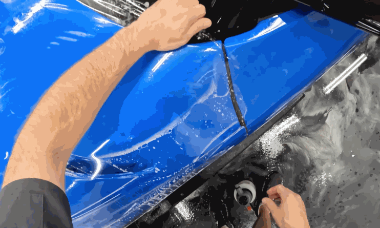 Hands stretching and positioning a clear paint protection film on the side panel of a blue car, with water and tools visible on the floor.