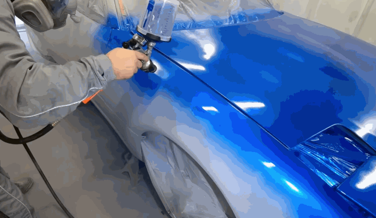 A technician in protective gear spray-paints a car with a vibrant blue color, covering the fender with smooth, even strokes.
