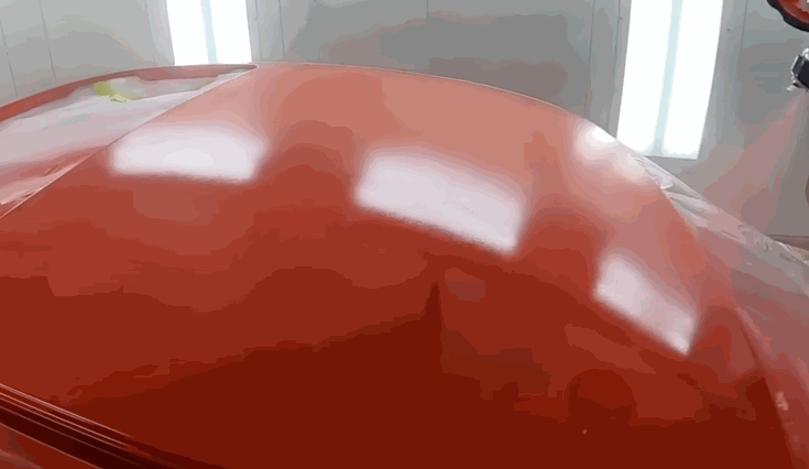 A fresh coat of glossy red paint is applied to a car hood inside a spray booth, reflecting overhead lights in a smooth, mirror-like finish.