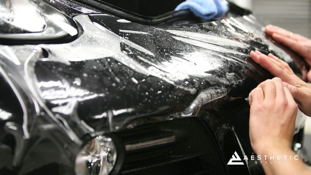 Close-up of a black car’s front end as a technician applies paint protection film, smoothing it over the glossy surface with a blue microfiber towel nearby.