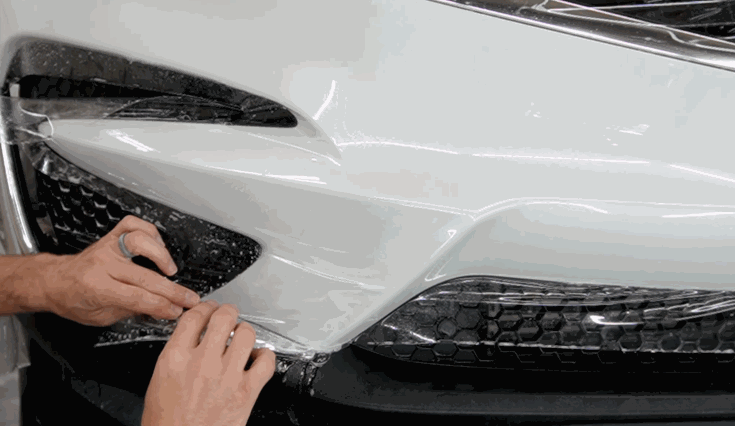 A technician carefully stretches and applies paint protection film to the full front corner of a white vehicle, covering the bumper and headlights.