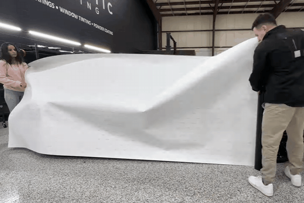 Two people carefully apply a large sheet of colored paint protection film (PPF) over a vehicle inside a detailing shop. The image captures the beginning stage of installation, emphasizing the scale and precision involved in the PPF application process.