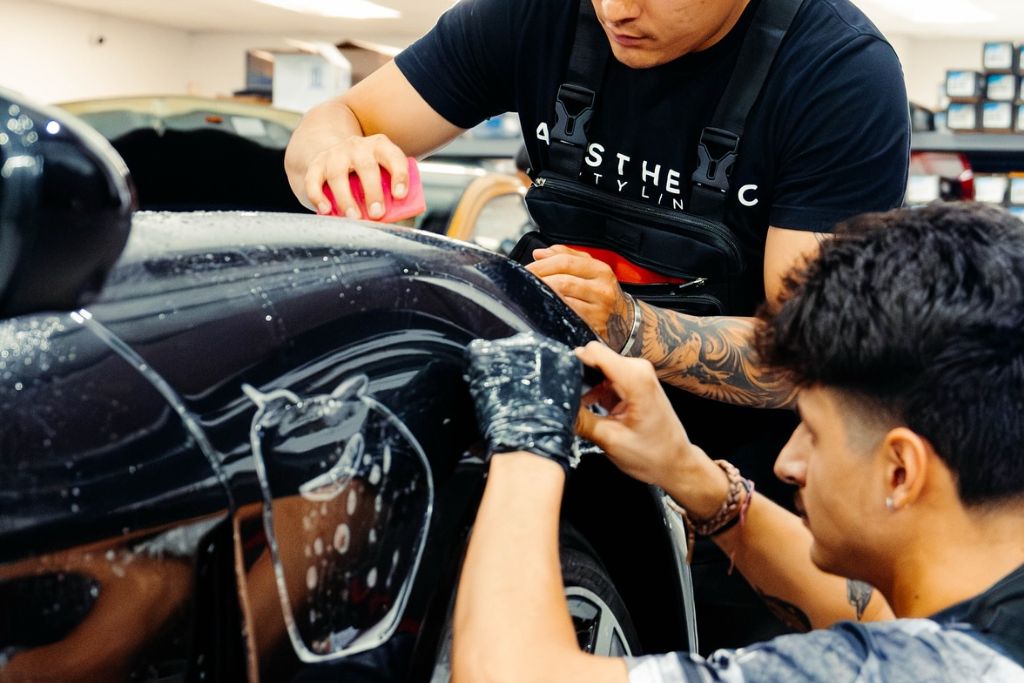 Two professional installers from Aesthetic Styling apply paint protection film to a glossy black car, using tools and precision techniques to ensure a smooth, bubble-free finish. The car's surface is wet, showing the careful process of aligning and securing the PPF.