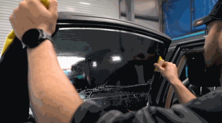 A technician uses a squeegee and yellow cloth while applying window tint to a large front side window of a vehicle inside a garage.