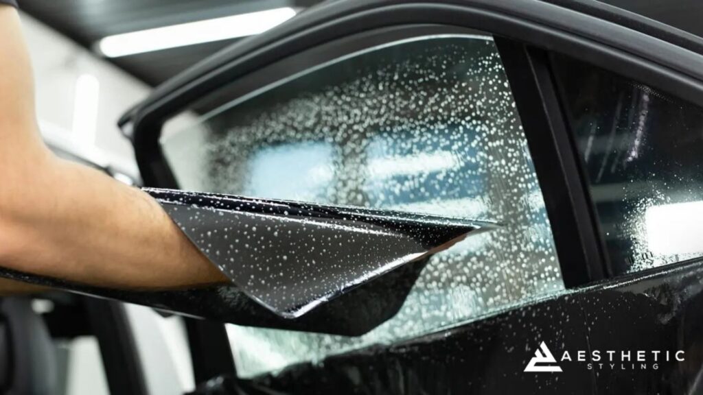 Close-up of a window tint film being positioned onto a soapy car window, with visible bubbles and the Aesthetic Styling logo in the corner.