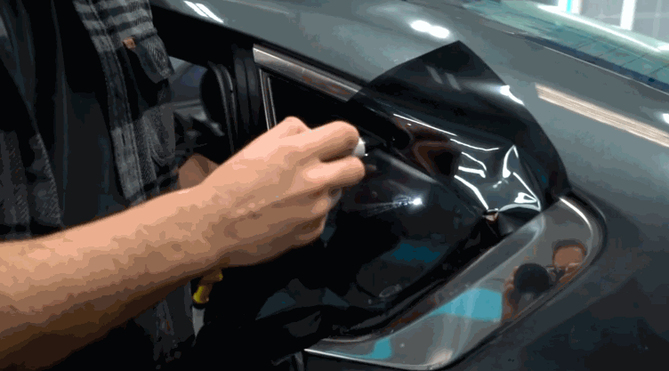 A person applies a dark window tint film to a car’s rear door glass, using a tool to smooth out the material for proper adhesion.