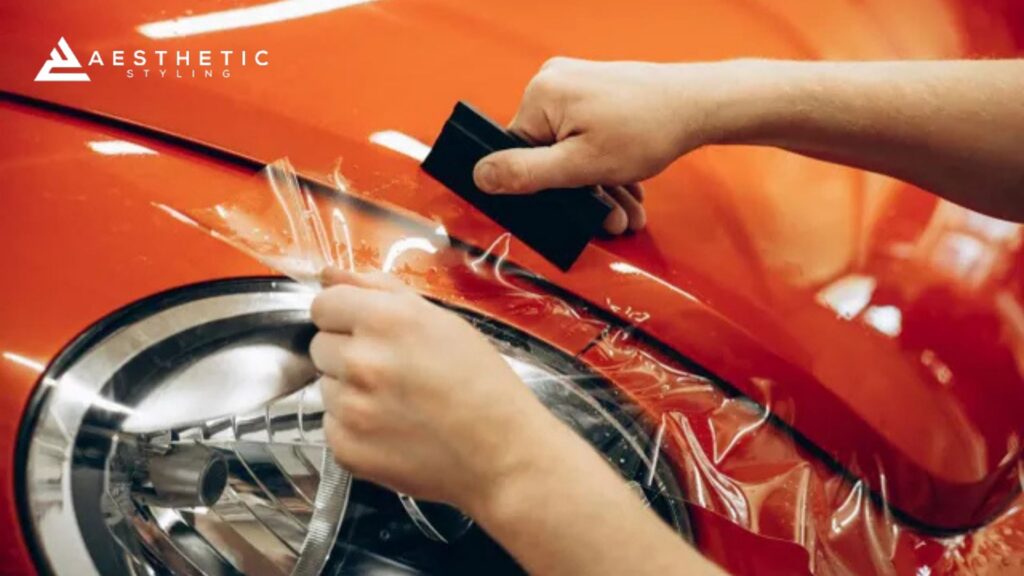 A person uses a black squeegee tool to smooth out paint protection film over the curved surface of a red car near the headlight, while another hand holds the film taut.