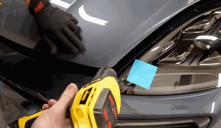 A technician uses a heat gun to apply paint protection film on the edge of a car hood, while another hand in black gloves holds the panel in place near the headlight.
