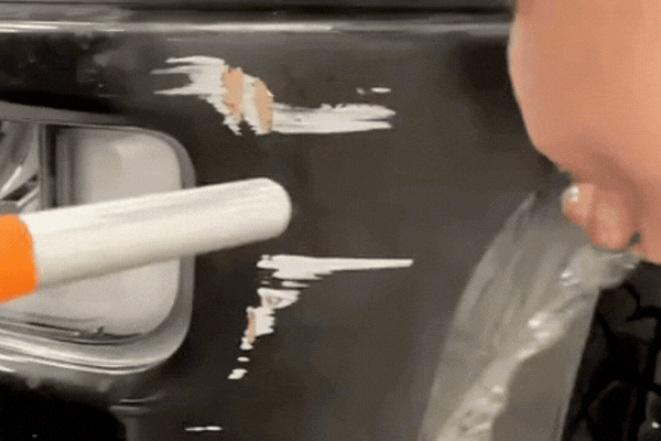 Close up of a black vehicle door with chipped paint and scratches near the handle, exposing lighter underlayers beneath the surface. A person holds a tool near the damaged area, highlighting how road salt, temperature shifts, and UV exposure can accelerate paint protection film deterioration in Naperville’s climate. The visible peeling and wear demonstrate common signs of clear bra damage caused by seasonal extremes.