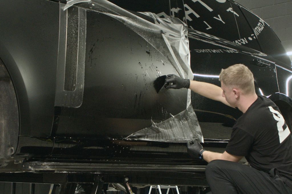 A technician wearing black gloves carefully removes aging paint protection film from the side panel of a black SUV inside a professional auto shop. The lifted film reveals moisture and surface marks underneath, showing why a professional PPF inspection in Naperville is essential to assess adhesive integrity, edge sealing, and topcoat condition. The image emphasizes the importance of safe removal to prevent paint damage on aging PPF.
